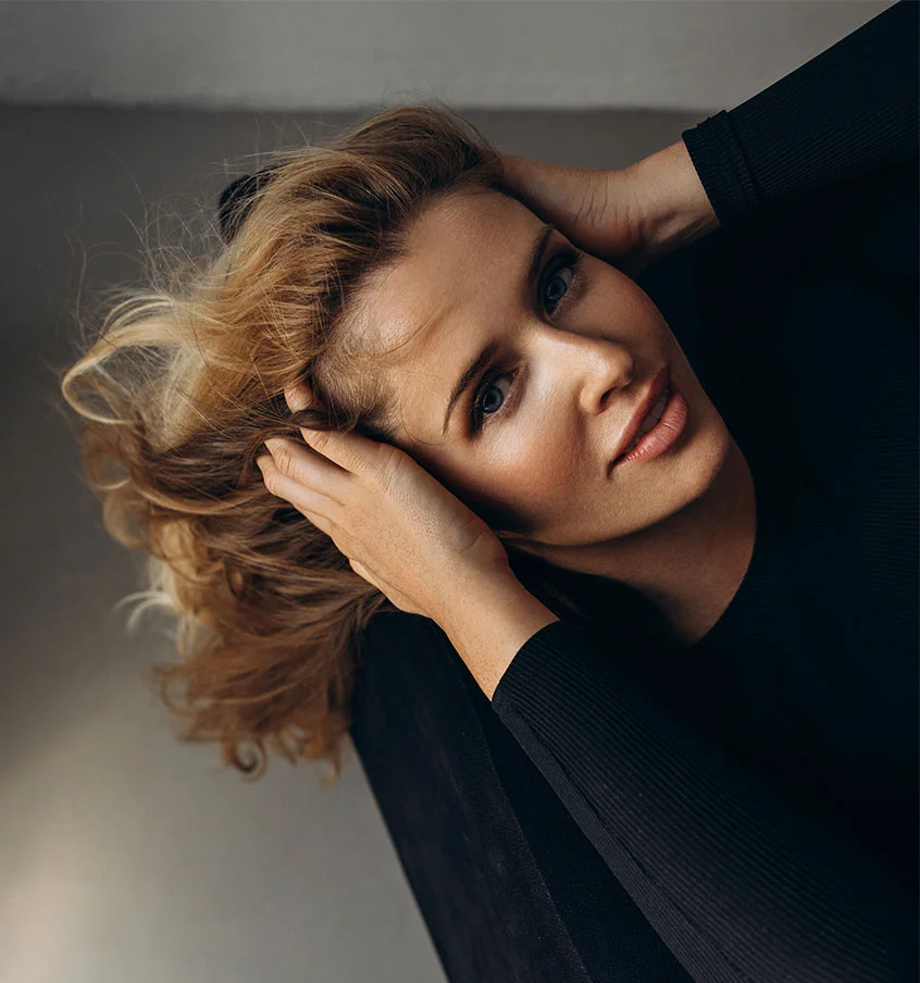 Woman in a black long sleeve, sitting in a chair, leaning her head back with her hands in her hair - Brow Lift in New York, NY