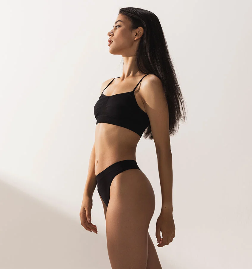 Woman with long dark hair, wearing a matching black underwear set, staning to the side against a minimal backdrop -Cellulite Reduction in New York, NY